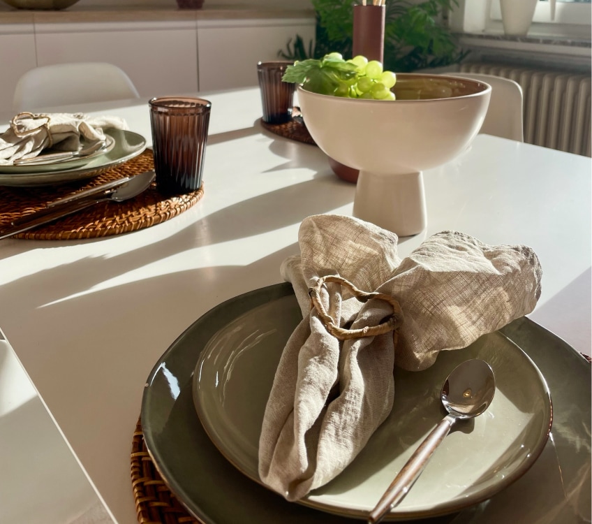 Ein weißer, gedeckter Esstisch mit grünen Tellern, braunen Gläsern und einer Schale mit Weintrauben.