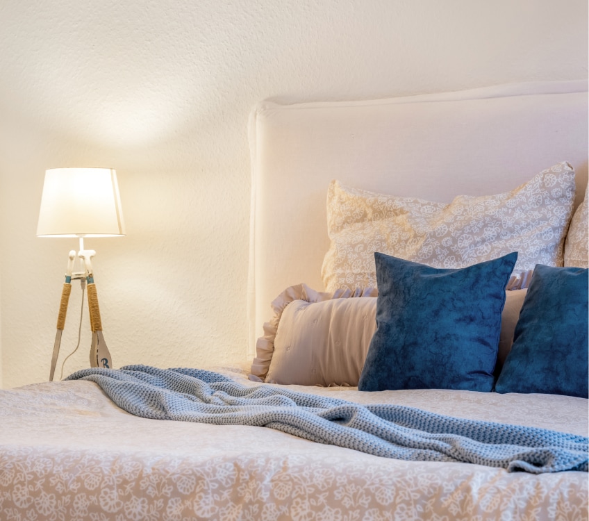 Stilvolles, weißes Schlafzimmer mit Bett, einer Stehlampe und blauen Kissen als Akzentfarbe. Umgesetzt von TraumStil Home Staging.